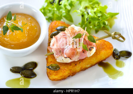 Roasted goat cheese wrapped in smoked bacon and served on crispy hash browns with fried pumpkin seeds, salad and fig mustard Stock Photo