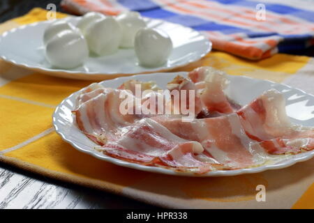 Raw bacon + Italian cherry-sized buffalo mozzarella cheese in background on colorful kitchen cloths lying on dark wooden table - Selective focus Stock Photo