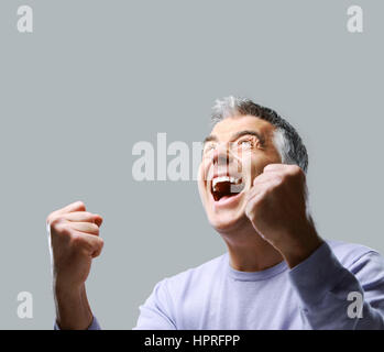 Portrait of excited mature man, raising his fists on grey background. Stock Photo