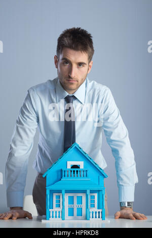Young real estate agent leaning on desk with model house. Stock Photo