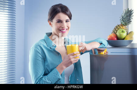 Young cheerful woman holding a yellow mug and leaning against refrigerator. Stock Photo