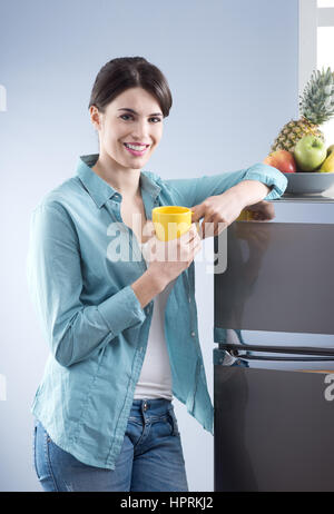 Young cheerful woman holding a yellow mug and leaning against refrigerator. Stock Photo