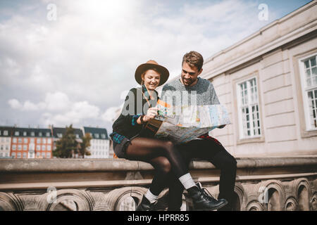 Young tourist couple exploring the navigation map of a city. Happy couple sitting on bridge on a cloudy day looking at a map. Stock Photo