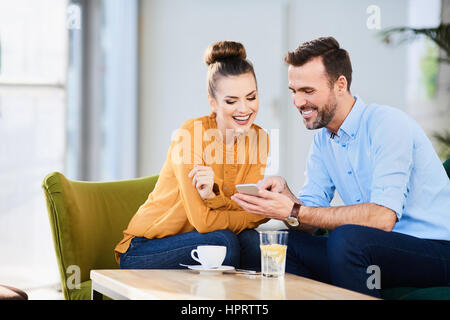 Young couple smiling and laughing as they check their mobile phone for text messages while sitting relaxing with drinks Stock Photo