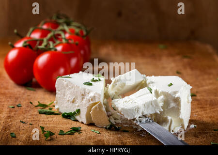 Feta cheese with knife and bunch of cherry tomatoes in background on wood with herbs Stock Photo