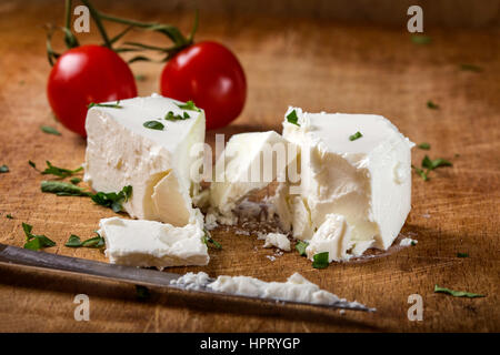 Feta cheese with knife and two cherry tomatoes in background on wood with herbs Stock Photo