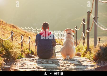 Man with dog on the trip in the mountains. Young tourist and his dog are resting and together watching the sunset. - back lit Stock Photo