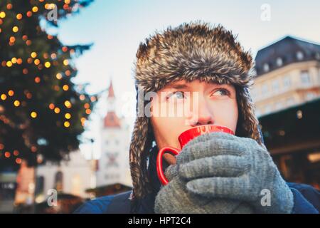 Young man drinking hot wine (or punch or tea or coffee) from red cup at the Christmas market - Munich, Germany Stock Photo