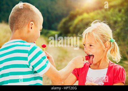 Children enjoying sunny day in holiday. Cheerful little boy and little girl tasting lollipops. Stock Photo