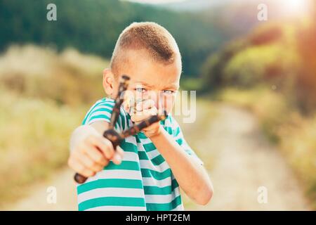 Naughty boy holding slingshot with stone. Little boy is playing in rural landscape. Stock Photo