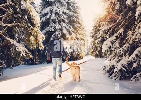 Trip to winter nature. Young man in warm clothes is walking with his labrador in snowy forest. Stock Photo