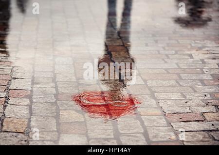 Rainy day. Reflection of young man with red umbrella in puddle on the city street during rain. Stock Photo