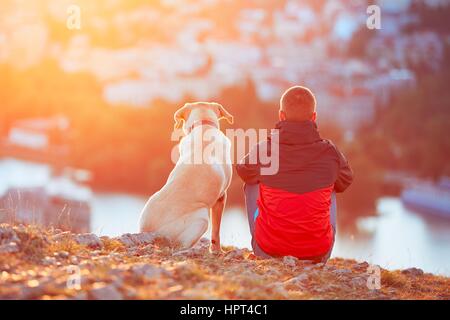 Enjoying sun. Pensive young man sitting on the hill with his dog (yellow labrador retriever). Amazing sunrise in the city. Prague in Czech Republic.