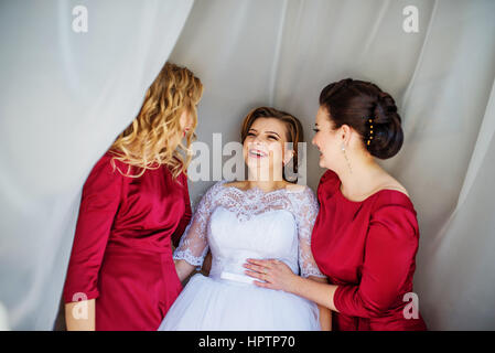 Cute blonde bride with bridesmaids posed on curtains at wedding morning day. Stock Photo