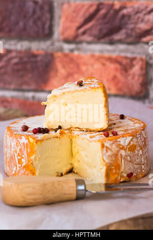 Soft cheese with cut off slice creamy texture, orange rind with mold, French, German, Alps, red pepper corns, close up, brick wall background Stock Photo