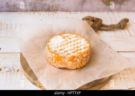 Whole wheel of soft French, German cheese with orange rind with mold on parchment paper, wood cutting board, concrete wall, rustic kitchen interior Stock Photo