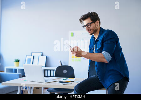 Designer sitting on desk in office using his smartphone Stock Photo