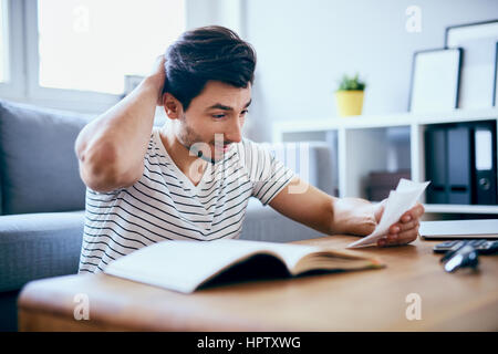 Worried man counting home finances sitting on floor in living room. Scratching his head. Stock Photo