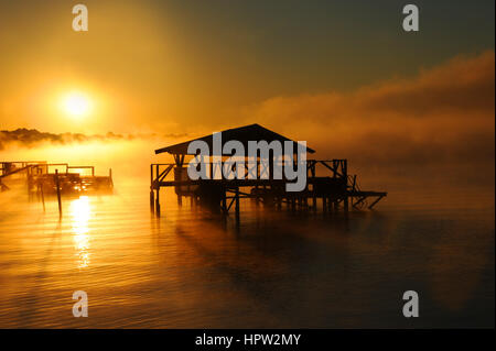 Early morning mist rises from Lake Chicot in Lake Village, Arkansas.  Wooden dock and boat house are silhouetted. Golden light covers lake.