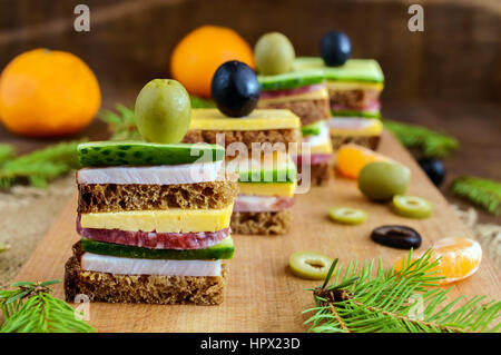 Mini sandwiches (canape) with fresh cucumbers, ham, cheese, olives, salami, black bread on a cutting board. Close up Stock Photo