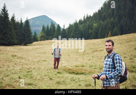 Portrait of two young men wearing backpacks and carrying trekking poles standing in a field while hiking in the hills Stock Photo