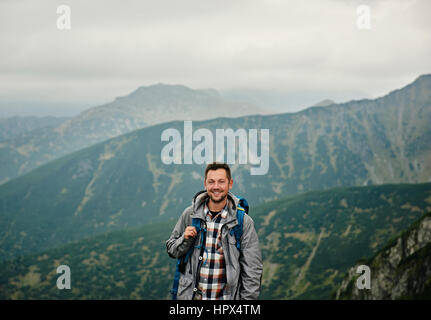 Man in hiking gear standing atop a mountain with a rolling landscape under an overcast sky behind him while out trekking in the wilderness Stock Photo