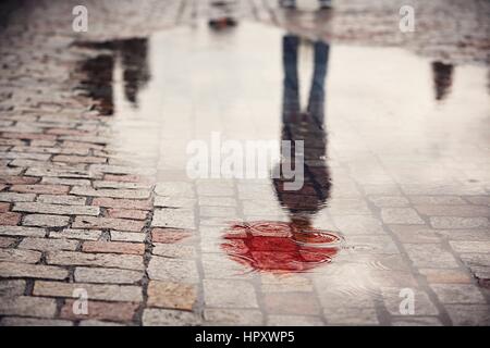 Rainy day. Reflection of young man with red umbrella in puddle on the city street during rain. Stock Photo