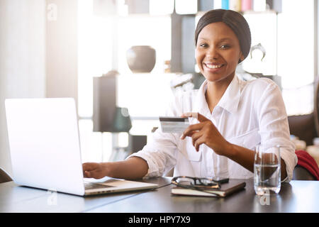 Good looking young black woman smiling at the camera while seated in front of her computer at her dining room table behind her notebook where she is m Stock Photo