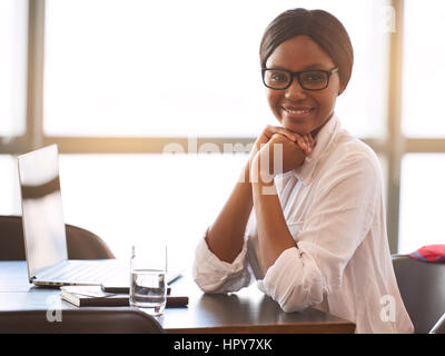 Young black woman wearing glasses looking at camera with a big smile resting her head on her hands while leaning on the desk with her elbows and large Stock Photo