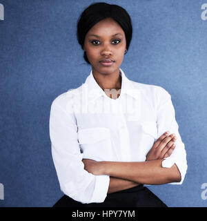 Confident young black woman looking at camera with a serious facial expression and her arms crossed while wearing a shite shirt, isolated against a bl Stock Photo
