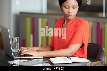 Well read adult black businesswoman busy typing while reading at the same time, while sitting in a colourful office space and wearing a bright orange  Stock Photo