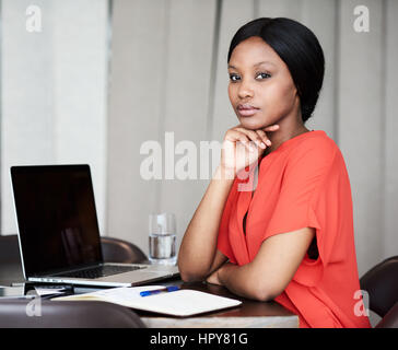 Attractive young adult businesswoman looking into the camera confidently while sitting at her desk in her work space while resting her head on her han Stock Photo