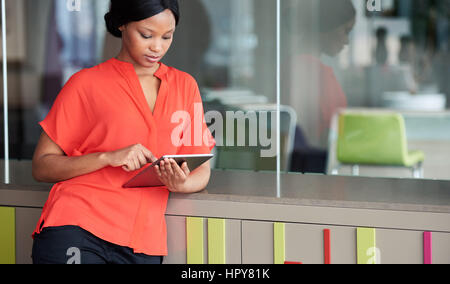 Modern young african american woman busy looking at the screen of the digital tablet that she is holding, while leaning against a colourful cabinet th Stock Photo