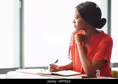 Young black businesswoman taking a little break from her work while looking out the window she is sitting next to with a pen still in her hands, while Stock Photo