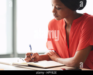 Beautiful young african american writer busy making hand written notes while wearing a bright orange blouse and sitting next to large bright windows. Stock Photo