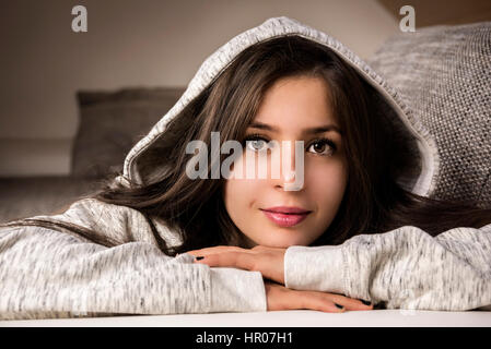 Beautiful, young brunette woman, relaxing on the couch, at home, dressed in a cozy hooded sweatshirt,  smiling at the camera. Stock Photo