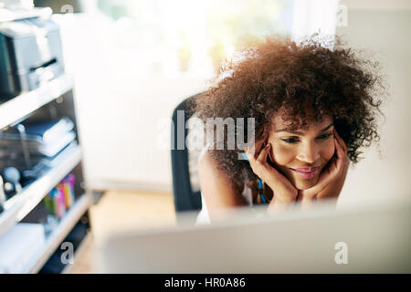 Young pretty afro-american woman looking at desk-top and smiling on blurred inside background. Stock Photo
