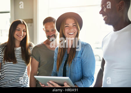 Four young stylish people smiling and having fun together while sharing tablet in light room. Stock Photo