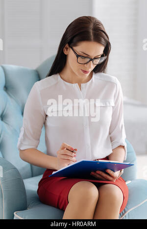 Developing a plan. Serious pleasant young business woman sitting in the armchair and taking notes while concentrating on her work Stock Photo
