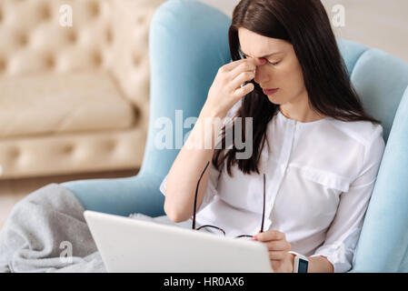 Need to rest. Unhappy tired brunette woman touching the bridge of her nose and holding glasses while suffering from the headache Stock Photo