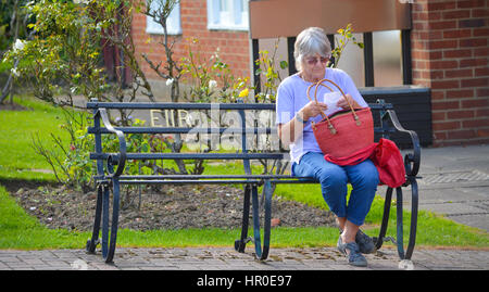 Mature woman sat on a bench in the summer sun, reading a shopping list and contemplating life. Stock Photo