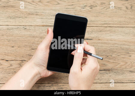 On old wooden table, one female hand holding a large smartphone, and the other writing on stylus it Stock Photo