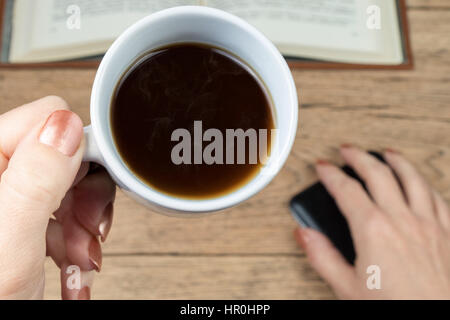 Female hand brings cup of hot coffee to camera close-up on a background of a wooden table with an open book Stock Photo