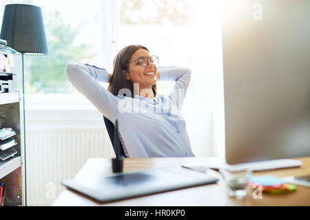 Happy single woman in long sleeve shirt and eyeglasses with arms folded behind head while seated at desk Stock Photo