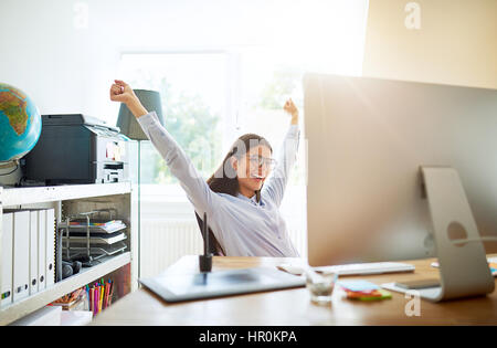 Single young excited woman celebrating something with both arms extended upward while seated at desk in small office Stock Photo