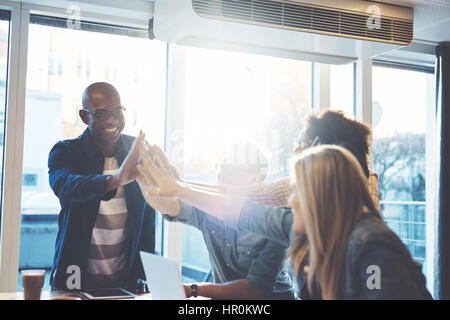 Young people in casual clothes giving high fives to each other as if celebrating something, against bright window in cafe or business office Stock Photo