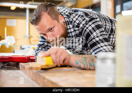 Craftsman using sanding paper on a guitar neck in a workshop for wood. Hard working man with tattoo and beard working with musical instruments. Stock Photo