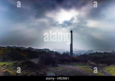A lone, tall brick industrial chimney stands silhouetted against a dramatic, overcast sky with sunbeams breaking through heavy clouds. Stock Photo