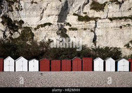 A row of white and red beach huts sit at the bottom of a chalk cliff on a pebble beach Stock Photo
