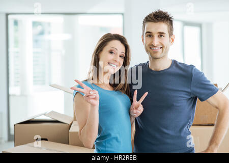 Loving couple posing in their new house surrounded by carton boxes, they are smiling at camera and she is making V signs Stock Photo